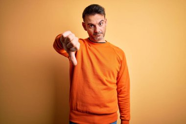 İzole edilmiş sarı arka plan üzerinde duran turuncu süveter giyen genç ve yakışıklı bir adam mutsuz ve kızgın görünüyor. Reddedilmiş ve olumsuz jestler yapıyor. Kötü ifade.