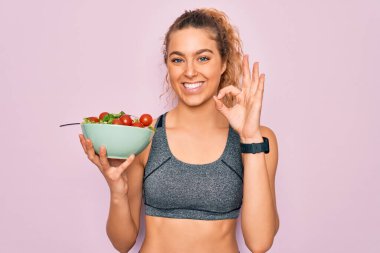 Genç, güzel, sarışın, mavi gözlü, sağlıklı salatalı bir kâse tutan sporcu kadın. Parmaklarıyla imzala, mükemmel bir sembol.