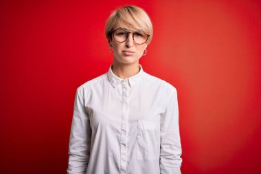 Kısa saçlı, kırmızı arka planda gözlük takan genç, sarışın iş kadını. Depresyona girmiş, üzgün ve korkmuş bir şekilde ağlıyor. Üzücü ifade.
