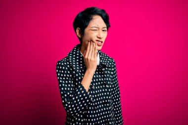 Genç ve güzel Asyalı bir kız günlük ceket giyiyor pembe arka planda duruyor diş ağrısı ya da diş hastalığından dolayı acı dolu bir ifadeyle ağıza dokunuyor. Dişçi