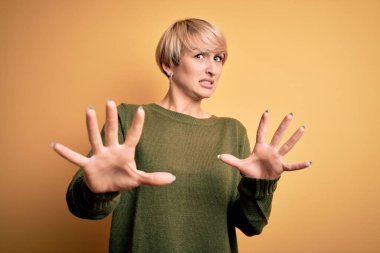 Modern kısa saçlı genç bir kadın sarı arka plan üzerine gündelik süveter giyiyor. Korkudan dehşete düşmüş ve korkmuş. El hareketlerini bırak, şok içinde bağır. Panik konsepti.