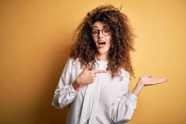 Kıvırcık saçlı ve piercingli genç ve güzel bir kadın. Gömlek ve gözlük takıyor. Kameraya hayret ediyor ve gülümsüyor..