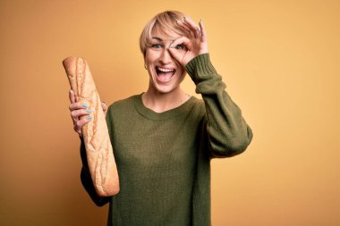 Sarışın, kısa saçlı genç bir kadın sarı arka planda taze ekmek ekmeği tutuyor. Gülümsüyor, tamam işaret veriyor, parmaklarından bakıyor.