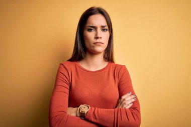 Sarı arka plan kuşkucu ve gergin, kolları çaprazlanmış, yüzü onaylamayan, sıradan bir tişört giyen genç, güzel esmer kadın. Negatif kişi.