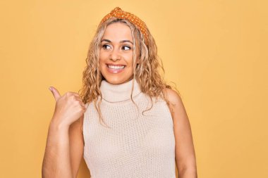 Güzel sarışın kadın, günlük tişört giyiyor ve sarı arka plan üzerinde dikdörtgen çiziyor, gülümsüyor ve başparmağıyla yan tarafa bakıyor..