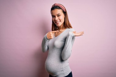 Genç, güzel bir kız hamile, pembe arka planda bebek bekliyor. Kameraya bakıp, el ve parmak işareti yaparken hayrete düşmüş ve gülümsüyor..