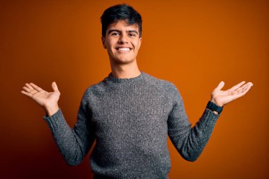 İzole edilmiş turuncu arka plan üzerinde duran sıradan süveter giyen genç yakışıklı adam gülümseyerek avuçlarını açıyor, sunuyor ve reklam karşılaştırması yapıyor ve dengeyi sağlıyor.