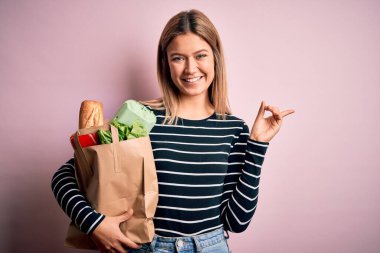 Genç ve güzel bir kadın, pembe arka plan üzerinden alışveriş torbası taşıyor. Elleri ve parmakları kenarı işaret ederek çok mutlu.