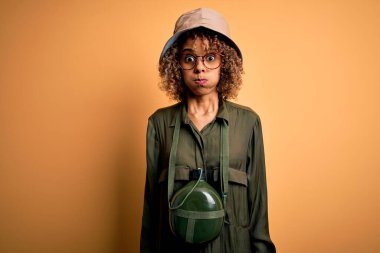 Genç Afro-Amerikan turist kadın tatilde. Kaşif şapkası ve matara takıyor. Komik suratıyla yanakları şişiyor. Havayla şişmiş ağız, çılgın bir ifade..