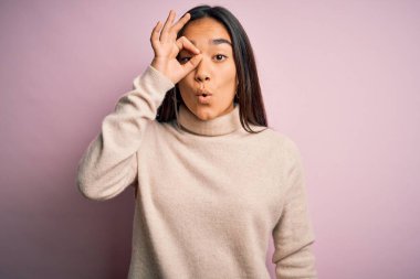Genç, güzel Asyalı kadın pembe arka planda balıkçı yaka kazak giyiyor. Şaşırmış yüz ifadesiyle şaşırmış, parmaklarından bakan gözler. İnançsız ifade.