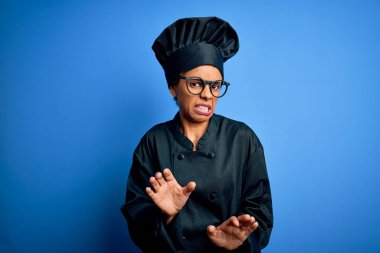 Afro-Amerikan aşçı kadın aşçı üniforması ve mavi arka planda şapka takıyor. Tiksindirici tepki verdiği için tiksinti ve tiksinti ifadesi takınıyor..