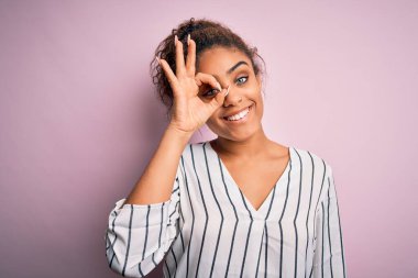 Genç ve güzel Afro-Amerikalı kız pembe arka planda duran çizgili tişört giyiyor. El hareketleriyle gülümsüyor, gözleriyle mutlu yüzüyle parmaklarının arasından bakıyor..