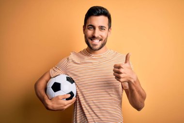 Sarı arka planda futbol topu tutan sakallı yakışıklı oyuncu elle mutlu bir jest yapıyor. Kameraya bakıp başarıyı gösteren ifade onaylanıyor.