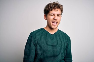 Sarışın, kıvırcık saçlı, beyaz arka planda yeşil kazak giyen, seksi bir ifadeyle kameraya bakan, neşeli ve mutlu yüzlü genç bir adam..