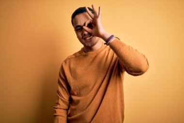 İzole edilmiş sarı arka planda dikilen sıradan bir süveter giyen genç Brezilyalı adam el hareketleriyle gülümsüyor ve mutlu yüzle parmaklarının arasından bakıyor..