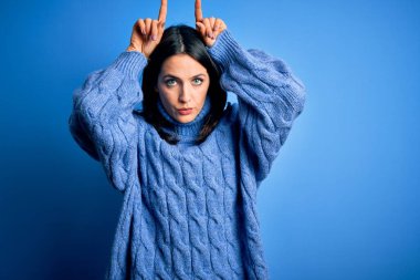 Mavi gözlü genç esmer kadın balıkçı yaka kazak giyiyor. Boğanın boynuzları gibi parmağıyla komik hareketler yapıyor.