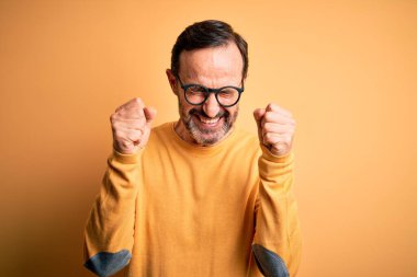 Kolları havada ve gözleri kapalı zaferi kutlarken gülümseyen orta yaşlı, ağarmış, günlük süveter ve gözlük takan, izole edilmiş sarı arka plan. Kazanan konsept.