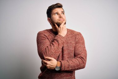 Sakallı ve mavi gözlü genç bir adam beyaz arka planda sıradan bir süveter giyiyor ve elleri çenesinde soru sormayı düşünüyor, düşünceli bir ifade. Düşünceli bir yüzle gülümsüyor. Şüphe kavramı.