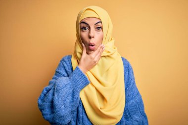 Genç, güzel, esmer, Müslüman kadın izole edilmiş sarı arka plan üzerine Arap başörtüsü takıyor. İnançsızlığa, şaşkınlığa ve elleri çenesinde olan şaşkınlık ifadesine hayran bakıyor.