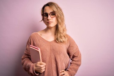 Genç, sarışın, gözlüklü, pembe arka planda not defteri tutan zeki yüzlü, ciddi düşünen güzel bir kadın.