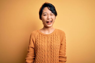 Genç, güzel Asyalı bir kız, günlük süveter ve Diadem giyip sarı arka planda dikiliyor, kameraya seksi bir ifadeyle bakıyor, neşeli ve mutlu bir yüzle..