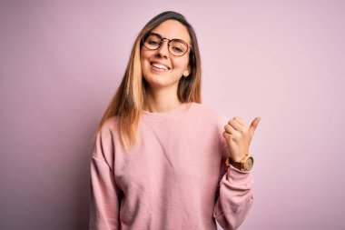 Mavi gözlü, pembe arka planda süveter ve gözlük takan, gülümseyen, gülümseyen ve baş parmağıyla yüzünü gösteren güzel sarışın kadın..