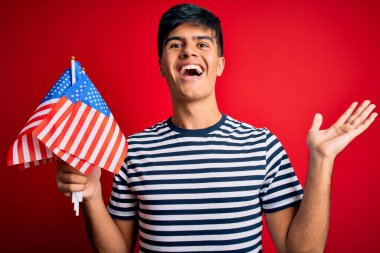 Amerika Birleşik Devletleri bayraklarını tutan genç, yakışıklı, vatansever adam bağımsızlık gününü kutluyor çok mutlu ve heyecanlı, kazanan ifade zaferi kutluyor büyük bir gülümsemeyle bağırıyor ve ellerini kaldırıyor.