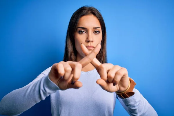 年轻美丽的黑发女人穿着休闲装站在蓝色背景上 用手指交叉表示拒绝 做阴性手势 — 图库照片