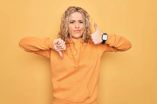 黄色の背景にカジュアルなスウェットシャツを着た若い美しいブロンドのスポーティな女性親指を上下に 意見の相違や合意表現を行います 狂気の葛藤 — ストック写真