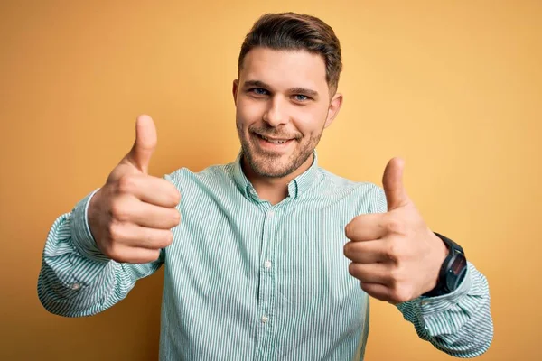 黄色の背景の上にエレガントな緑のシャツを着て青い目を持つ若いビジネスマンは 手で積極的なジェスチャーを行うことを承認し 親指を笑顔と成功のために幸せ 勝者のジェスチャー — ストック写真