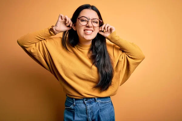 若いブルネットの女性は黄色の孤立した背景に眼鏡とカジュアルなセーターを着て笑顔指で耳を引く 面白いジェスチャー オーディション問題 — ストック写真