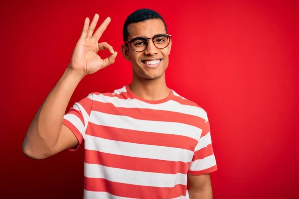 若いハンサムなアフリカ系アメリカ人の男性が手と指でOkの署名を行う肯定的な笑みを浮かべてカジュアルなストライプのTシャツとメガネを着ている 成功した表現 — ストック写真