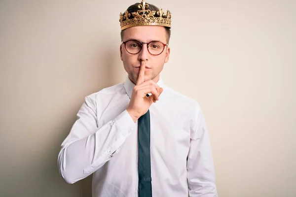 Young handsome caucasian business man wearing golden crown over isolated background asking to be quiet with finger on lips. Silence and secret concept.