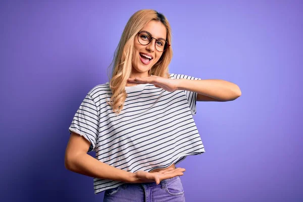 年轻美丽的金发女人 穿着条纹T恤 戴着眼镜 背景是紫色的 手握着大号和大号的标志 是尺寸的象征 笑着看着摄像机 计量概念 — 图库照片