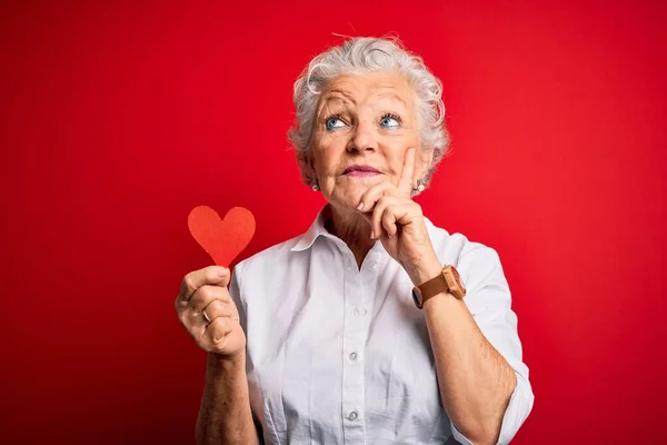Senior beautiful woman holding paper heart standing over isolated red background serious face thinking about question, very confused idea