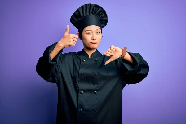 Young beautiful chinese chef woman wearing cooker uniform and hat over purple background Doing thumbs up and down, disagreement and agreement expression. Crazy conflict
