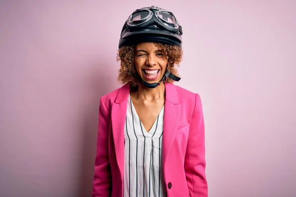 アフリカ系アメリカ人オートバイの女性とともに巻き毛を身に着けています本ヘルメット上のピンクの背景ウィンク見ますカメラでセクシー式 陽気で幸せな顔 — ストック写真
