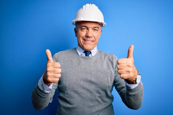 Мужчина средних лет с седыми волосами, одетый в защитный шлем на голубом фоне знак успеха делает позитивный жест рукой, большие пальцы вверх улыбается и счастлив. Веселое выражение лица и жест победителя
.