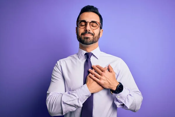 Handsome businessman with beard wearing casual tie and glasses over purple background smiling with hands on chest with closed eyes and grateful gesture on face. Health concept.