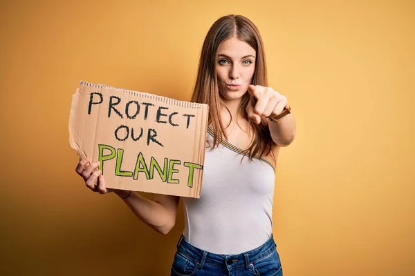 Young redhead woman asking for enviroment holding banner with protect planet message pointing with finger to the camera and to you, hand sign, positive and confident gesture from the front