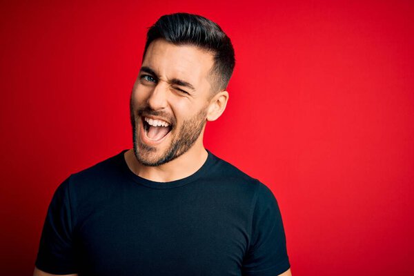 Young handsome man wearing casual black t-shirt standing over isolated red background winking looking at the camera with sexy expression, cheerful and happy face.