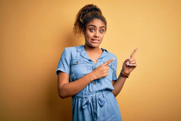 Young beautiful african american girl wearing denim dress standing over yellow background Pointing aside worried and nervous with both hands, concerned and surprised expression
