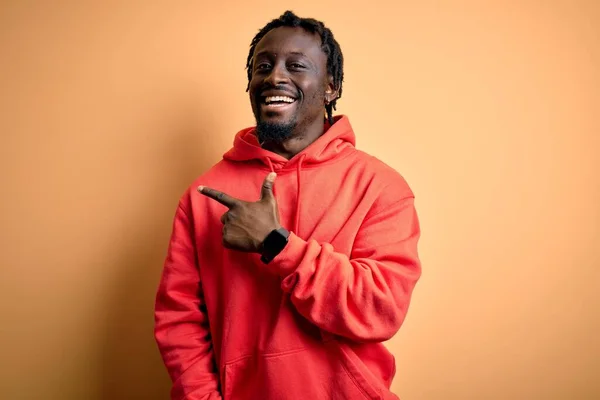 Young african american sporty man wearing sweatshirt with hoodie over yellow background cheerful with a smile of face pointing with hand and finger up to the side with happy and natural expression on face