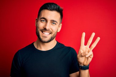 Siyah bir tişört giyen yakışıklı genç adam izole edilmiş kırmızı arka planın üzerinde duruyor ve üç numaralı parmaklarıyla gülümseyerek kendinden emin ve mutlu bir şekilde onu işaret ediyor..