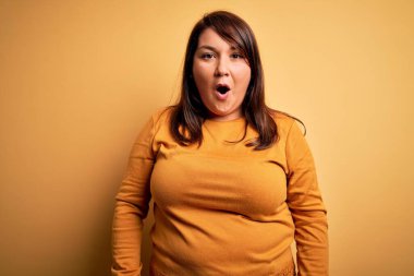 Güzel esmer ve cüsseli bir kadın izole edilmiş sarı arka plan üzerine gündelik bir kazak giyip şaşkınlık ve şaşkınlık içinde ifade, korku ve heyecan dolu yüz ifadesiyle şoka girmiş..