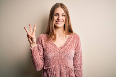 İzole edilmiş beyaz arka planda pembe kazak giyen genç, güzel, kızıl saçlı kadın üç numaralı parmaklarıyla gülümseyerek kendinden emin ve mutlu bir şekilde onu işaret ediyor..
