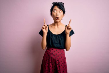 Genç, güzel Asyalı bir kız, günlük elbiseler giyip pembe arka planda dikilmiş şaşkın şaşkın bakıyor, parmaklarıyla işaret ediyor ve kollarını kaldırıyordu..