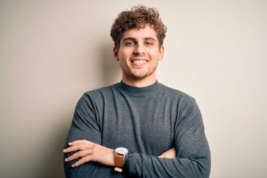 Sarışın, kıvırcık saçlı, beyaz arka planda rahat süveter giyen, kollarını kavuşturup kameraya bakan genç, yakışıklı bir adam. Pozitif kişi.