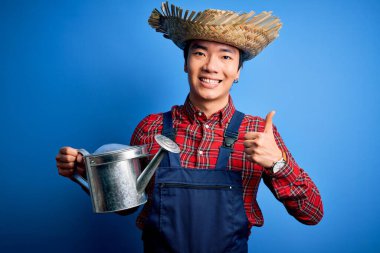 Önlük ve hasır şapka takan genç, yakışıklı çiftçi elinde büyük bir gülümsemeyle mutlu. Tamam işareti yap, başparmağını parmaklarınla kaldır, mükemmel işaret.