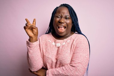 Afro-Amerikalı, örgü örgülü, pembe arka planda sıradan bir süveter giyen ve kameraya gülümseyen, zafer işareti yapan iri yarı bir kadın. İki numara..